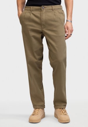 Homme portant un pantalon plissé vert olive avec poches latérales, des baskets beiges et une chemise noire, mains détendues le long du corps sur fond uni.