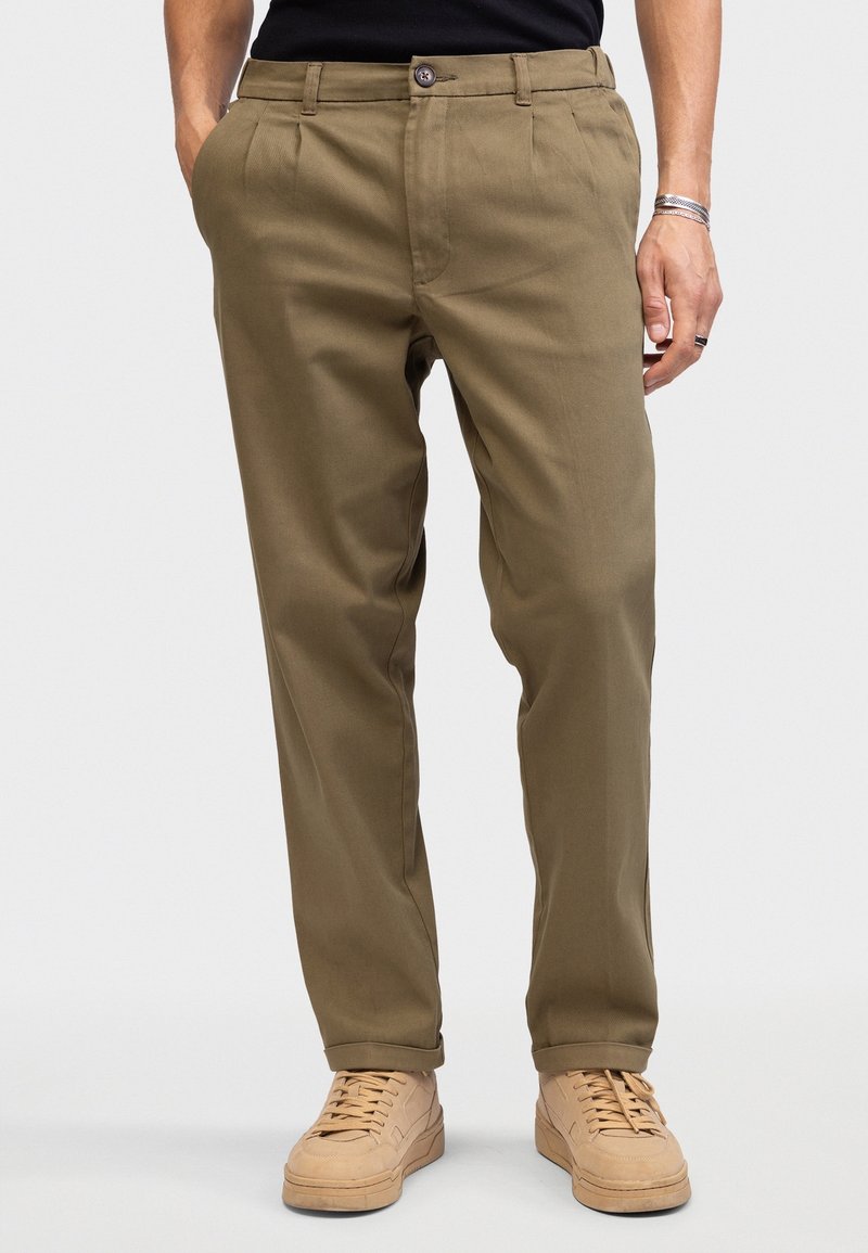 Homme portant un pantalon plissé vert olive avec poches latérales, des baskets beiges et une chemise noire, mains détendues le long du corps sur fond uni.