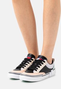 Sneaker nere e beige con suola bianca, dotate di tomaia in pelle liscia, lacci neri e un piccolo logo rosso e blu.