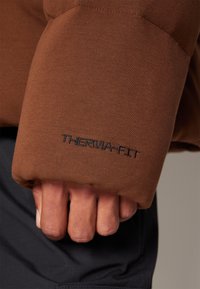 Manica di giacca Terma-Fit marrone con texture liscia; presenta un logo ricamato nero; indossata da una mano in un indumento nero sottostante.