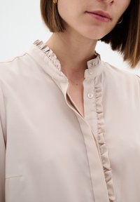 Blusa beige con colletto arricciato e patta frontale, dotata di una chiusura a un solo bottone e tessuto liscia.