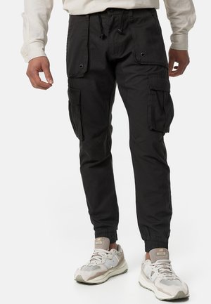 Mann trägt schwarze Cargohosen mit mehreren Taschen und weiße Sneaker, steht vor einem schlichten Hintergrund.