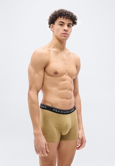 Άνδρας χωρίς πουκάμισο με σγουρά μαλλιά, φορώντας μπεζ μποξεράκια Polo Ralph Lauren, στέκεται μπροστά σε απλό ανοιχτό φόντο.