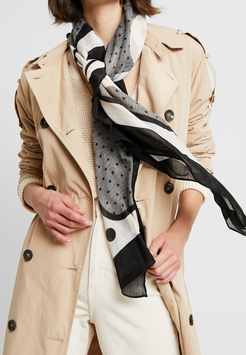 Femme portant un trench-coat beige, un pull crème, un pantalon blanc et une écharpe à motifs noir et blanc autour du cou.