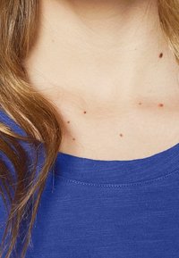 Modré tričko s kulatým výstřihem, vystavené na pleti s několika malými hnědými névy a měkkými, vlnitými vlasy částečně viditelnými.