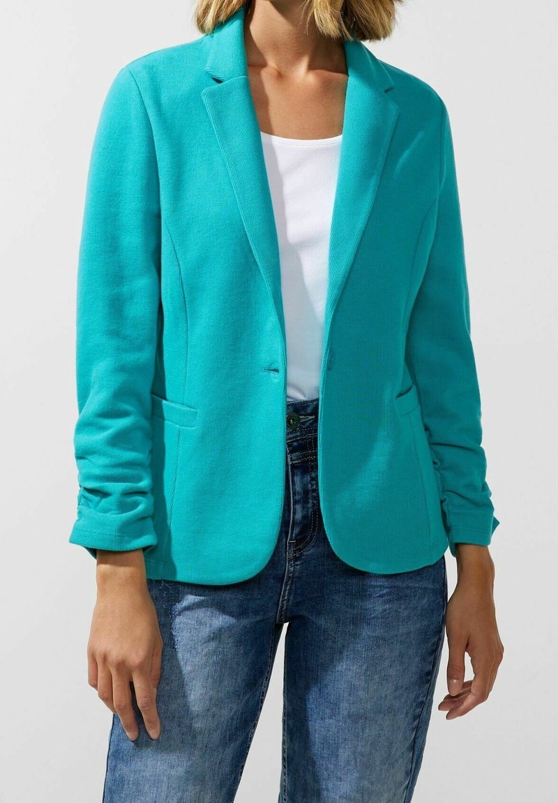 Blazer turquoise à revers crantés porté sur une chemise blanche, associé à un jean en denim bleu sur une personne devant un fond uni.