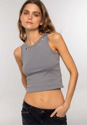 Joven mujer vestida con un crop top gris sin mangas con ojales metálicos cerca del escote y jeans negros con tachuelas, posando con una mano en el bolsillo.