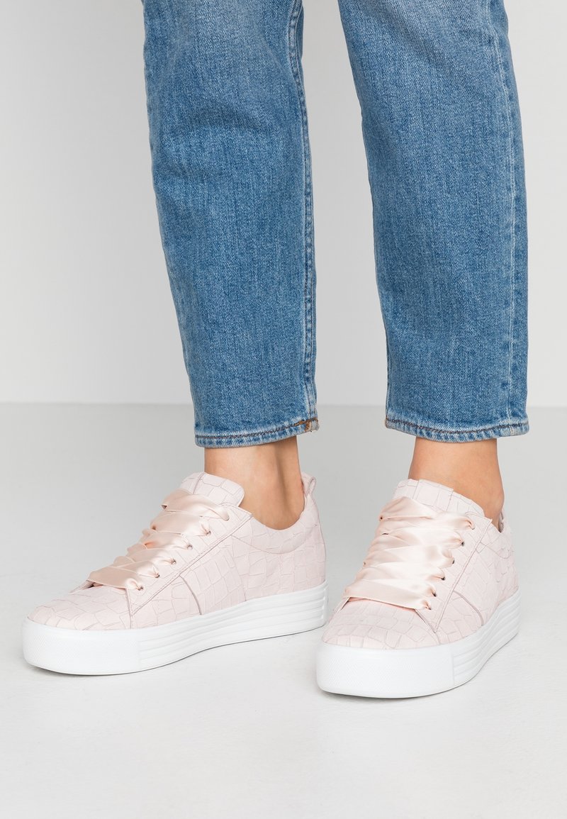 Baskets rose clair avec une texture crocodile, des lacets en satin et une épaisse semelle blanche. Associées à un jean bleu, mettant en avant un look décontracté.