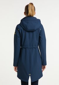 Námořnická modrá nepromokavá bunda s kapucí, stahovací pas a dvě zlaté akcenty na zádech. Hladká, texturovaná látka, délka ke kolenům.