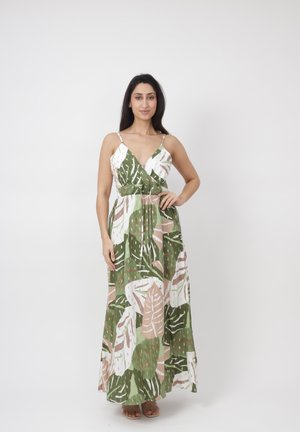 Kvinde med langt mørkt hår iført en ærmeløs maxi-kjole med grønne og beige tropiske bladprint, stående med den ene hånd på hoften.