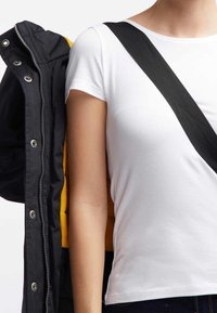 Vit kortärmad t-shirt som bärs under en svart jacka med knappar. En gul insida är synlig. Svart rem synlig över axeln.