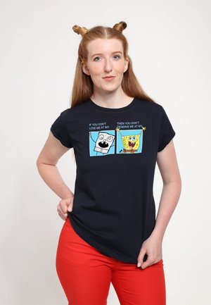 Jeune femme aux cheveux roux coiffés en deux chignons porte un t-shirt noir SpongeBob SquarePants et un pantalon rouge vif, debout devant un fond uni.