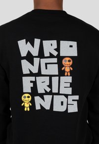 Zwarte sweatshirt met de tekst "WRONG FRIENDS" in grote grijze letters. Onder de tekst staan twee kleurrijke cartoonpoppen.
