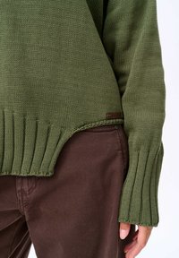 Wciągany sweter w oliwkowym kolorze z prążkowanymi mankietami i dołem, charakteryzujący się lekką teksturą oraz małą brązową metką logo blisko dolnej krawędzi.