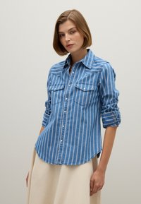 Donna dai capelli corti che indossa una camicia a righe blu e bianche con maniche arrotolate e una gonna beige, su uno sfondo neutro.
