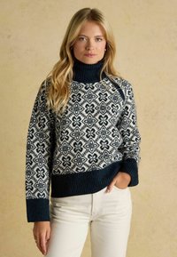 Strick-Turtleneck-Pullover in Marineblau und Creme mit floralen Mustern, gerippten Bündchen und einer lockeren Passform. Getragen über hellbeigen Hosen.