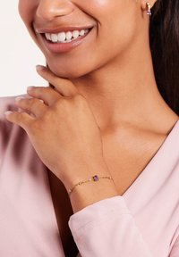 Gouden kettingarmband met een paarse edelsteen, gedragen om een pols. Glad van textuur en delicate vormgeving, aangevuld met een zacht roze top.