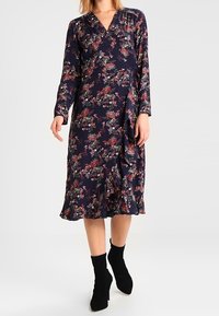 Marineblaue Blumenkleid mit langen Ärmeln, gerafftem Saum und lebhaften rosa und grünen Mustern. Aus leichtem Stoff gefertigt, kombiniert mit schwarzen Knöchelstiefeln.