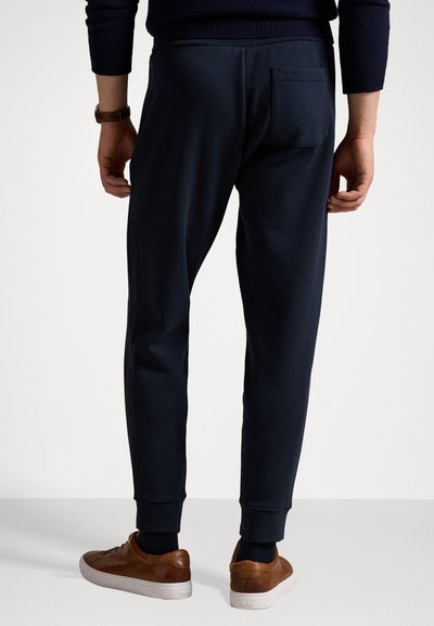 Marineblå joggebukser med smal passform, mykt stoff, elastisk midje og baklomme. Kombinert med brune lær-sneakers.