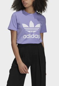 Lila bomulls t-shirt med en stor vit Adidas-logga och tre-randsdesign på framsidan, tillsammans med svarta vida byxor.