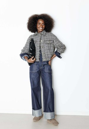 Femme avec une coiffure afro naturelle portant une veste texturée, un jean large retroussé, des mocassins et tenant une pochette noire.