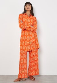 Orange gemusterte Tunika und Hose im Blattdesign, aus leichtem Stoff mit lockerer Passform. Kombiniert mit flachen Sandalen.