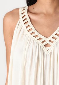 Top sans manches blanc avec un décolleté en V, ornée de détails en crochet et d'un tissu texturé qui tombe doucement.