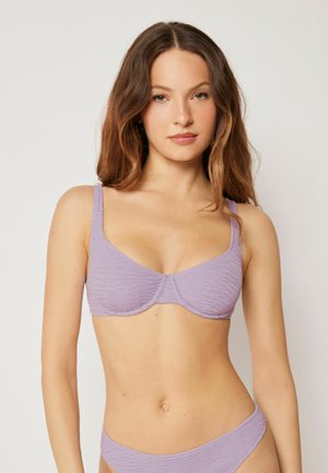 Junge Frau mit langen braunen Haaren, die einen strukturierten lavendelfarbenen Bikini-Oberteil und -Unterteil trägt und vor einem schlichten hellen Hintergrund steht.