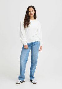 Weiße, gerippte Strickjacke mit langen, puffigen Ärmeln, kombiniert mit hellblauen geraden Jeans und schwarz-weißen Schuhen. Einfaches Design und Textur.