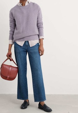 Personne portant un pull lavande par-dessus une chemise rayée, un jean bleu à jambes larges, des chaussures noires et tenant un sac à main en cuir marron.