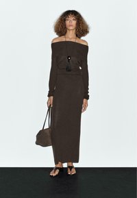 Frau mit lockigem Haar trägt ein schulterfreies, dunkelbraunes Kleid, eine schwarze Anhänger-Halskette, trägt eine braune Handtasche und schwarze Sandalen auf einem schwarzen Boden.