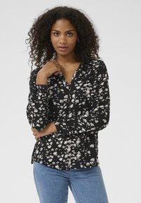 Blouse à manches longues noire avec un motif floral en blanc et beige, dotée d'un décolleté en V et de boutons sur les poignets.