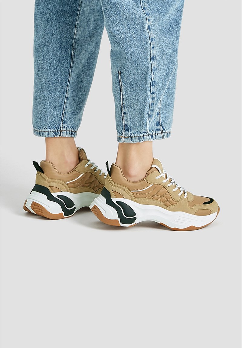 Baskets beiges et noires avec une semelle blanche épaisse, une tige texturée et des accents noirs. Elles comportent des lacets blancs et une languette de maintien au talon.