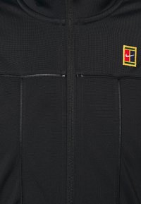 Chaqueta negra con cremallera y costuras verticales, y un parche rectangular colorido en el lado izquierdo del pecho.