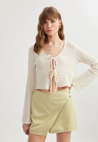 Suéter corto blanco con tejido texturizado, mangas largas y lazo frontal. Combinado con pantalones cortos de talle alto amarillos claros con detalles de botones.