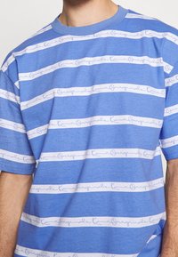 Blå bomullströja med horisontella vita ränder och handskriven textdesign. Rund hals och korta ärmar. Slät textur.