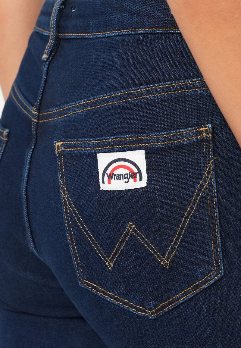 Donkerblauwe denim jeans met gele stiksels, een kenmerkend 'W'-ontwerp op de achterzak en een wit Wrangler-logo label.