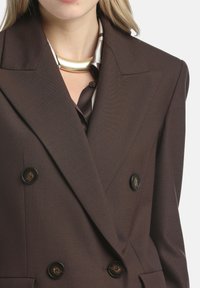 Blazer marrón de doble botonadura con una textura suave, amplios solapas y grandes botones negros. Complementado con una camisa de seda brillante debajo.