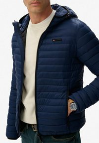 Mężczyzna w granatowej pikowanej kurtce z kapturem noszący białą sweter, z ręką w kieszeni, pokazujący brązowy skórzany zegarek na nadgarstku z okrągłą czarną tarczą.