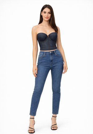 Kobieta stojąca, ubrana w ciemny denimowy top gorsetowy bez ramiączek, wysokowalne niebieskie dżinsy oraz czarne sandały na wysokim obcasie z paskami, na białym tle.