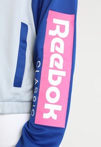 En blå och ljusgrå jackärm visar en rosa vertikal Reebok Classic-logotyp på det blå tyget.