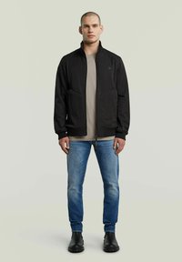 Svart zip-up jacka med sidofickor, bärd över en beige skjorta, tillsammans med blå jeans och svarta ankelstövlar. Slät textur, avslappnad design.