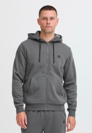Sudadera con cremallera gris hecha de una tela suave, que cuenta con un bolsillo frontal, capucha ajustable y un pequeño logo en el pecho. Textura suave.