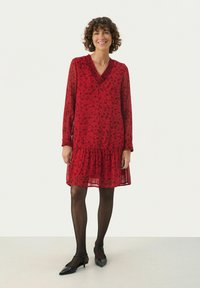 Rotes Blumenkleid mit langen Ärmeln, V-Ausschnitt und mehrlagigem Rüschensaum. Durchscheinender Stoff und feine Textur, kombiniert mit schwarzen spitzen Schuhen.