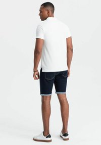 Vit kortärmad polo, mörka denimshorts med uppvikta fållar, och vita sneakers med svarta accenter. Utsikt bakifrån.