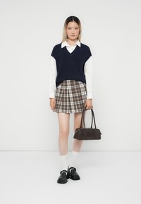 Pull marin en v sur une chemise blanche à col, associé à une jupe à carreaux beige et marron. Chaussures noires imposantes, chaussettes blanches et un sac à main foncé.