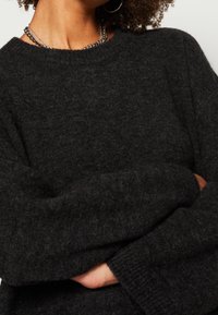Personne portant un pull en peluche gris foncé, un collier chaîne en argent et une boucle d'oreille anneau en argent, les bras croisés à la taille.