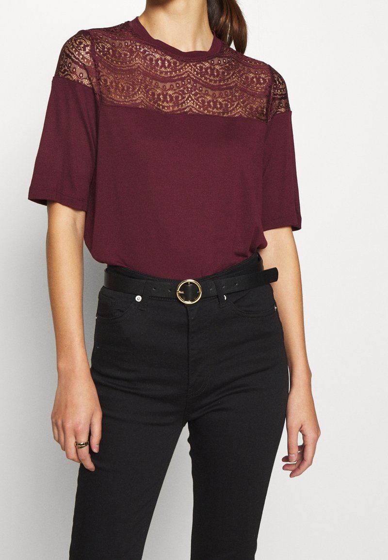 Femme portant un haut bordeaux à manches courtes avec un détail en dentelle sur le haut de la poitrine et un pantalon taille haute noir avec une ceinture noire.