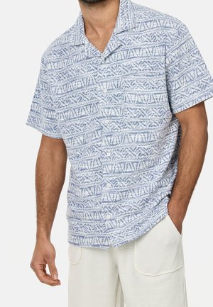Mężczyzna noszący koszulę z krótkim rękawem zapinaną na guziki z niebiesko-białymi geometrycznymi wzorami, w połączeniu z jasnymi, teksturowanymi spodniami.