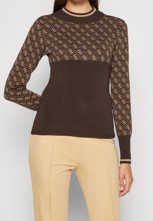 Frau trägt einen braunen Pullover mit langen Ärmeln und einem beigefarbenen geometrischen Muster auf der oberen Brust und den Ärmeln, kombiniert mit beigen Hosen.